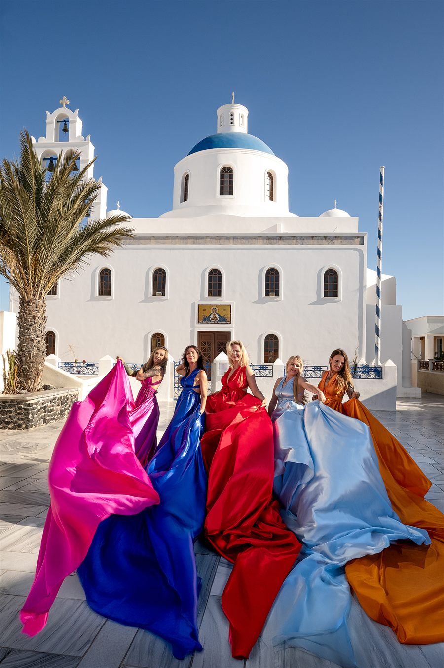 Kobiety w kolorowych sukniach przed kościołem na Santorini - każdy filar ma swój kolor